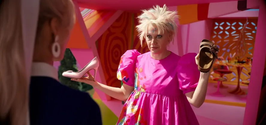 boneca barbie em live action segura um salto cor de rosa em uma mão e uma birkenstock bege de outra